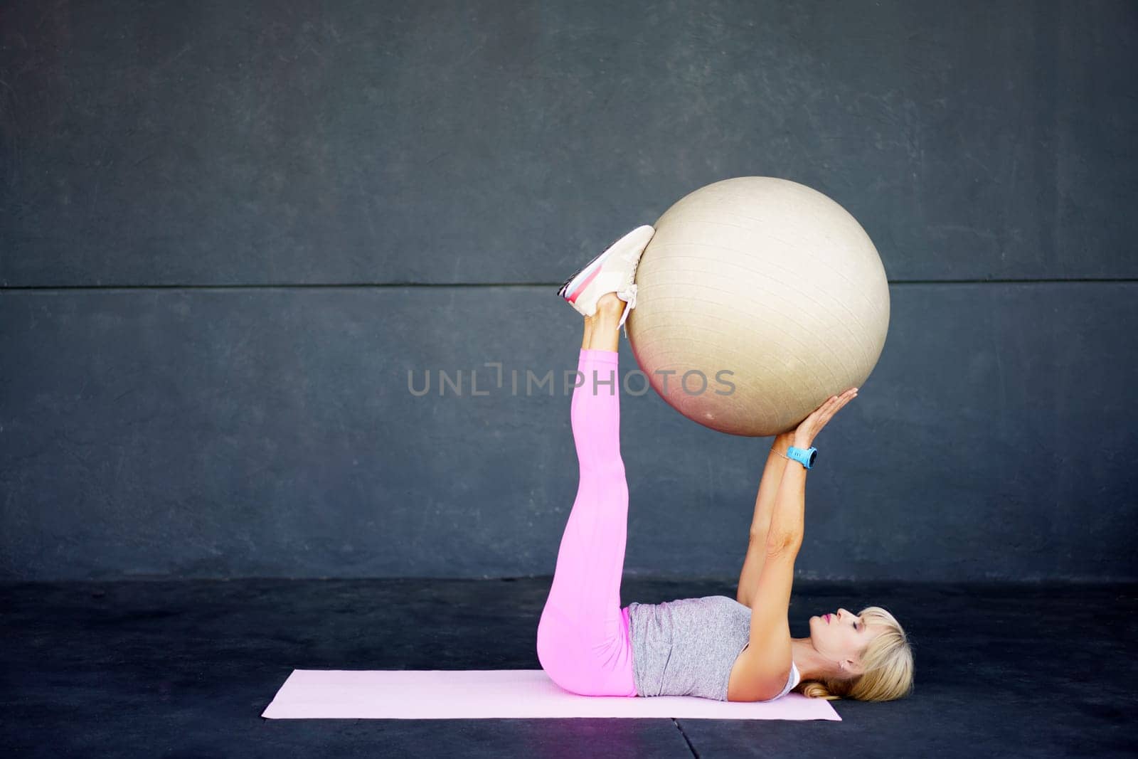 Side view of fit young female in activewear lying on mat, and doing exercise with fitness ball while practicing workout against gray wall at modern gym