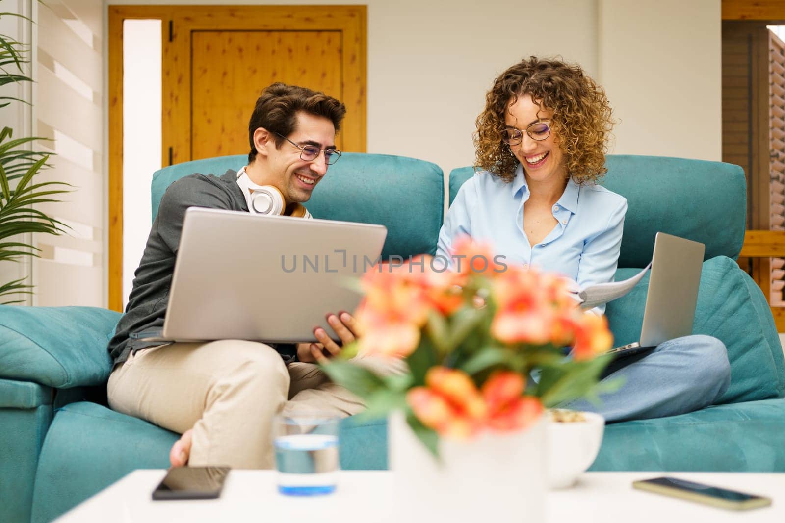 Positive young male and female freelancers, in casual clothes smiling and browsing laptop while sitting on sofa and working together on remote project in living room