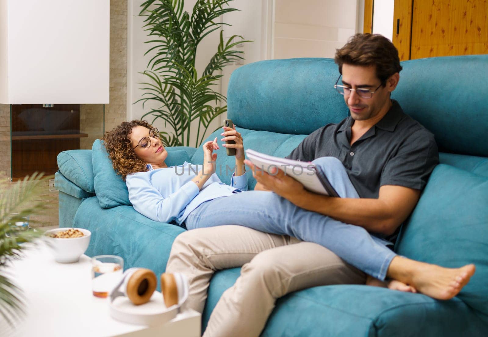 Happy young curly haired wife lying on back and browsing smartphone, while stretching legs on focused husband sitting and reading notebook over sofa in cozy living room