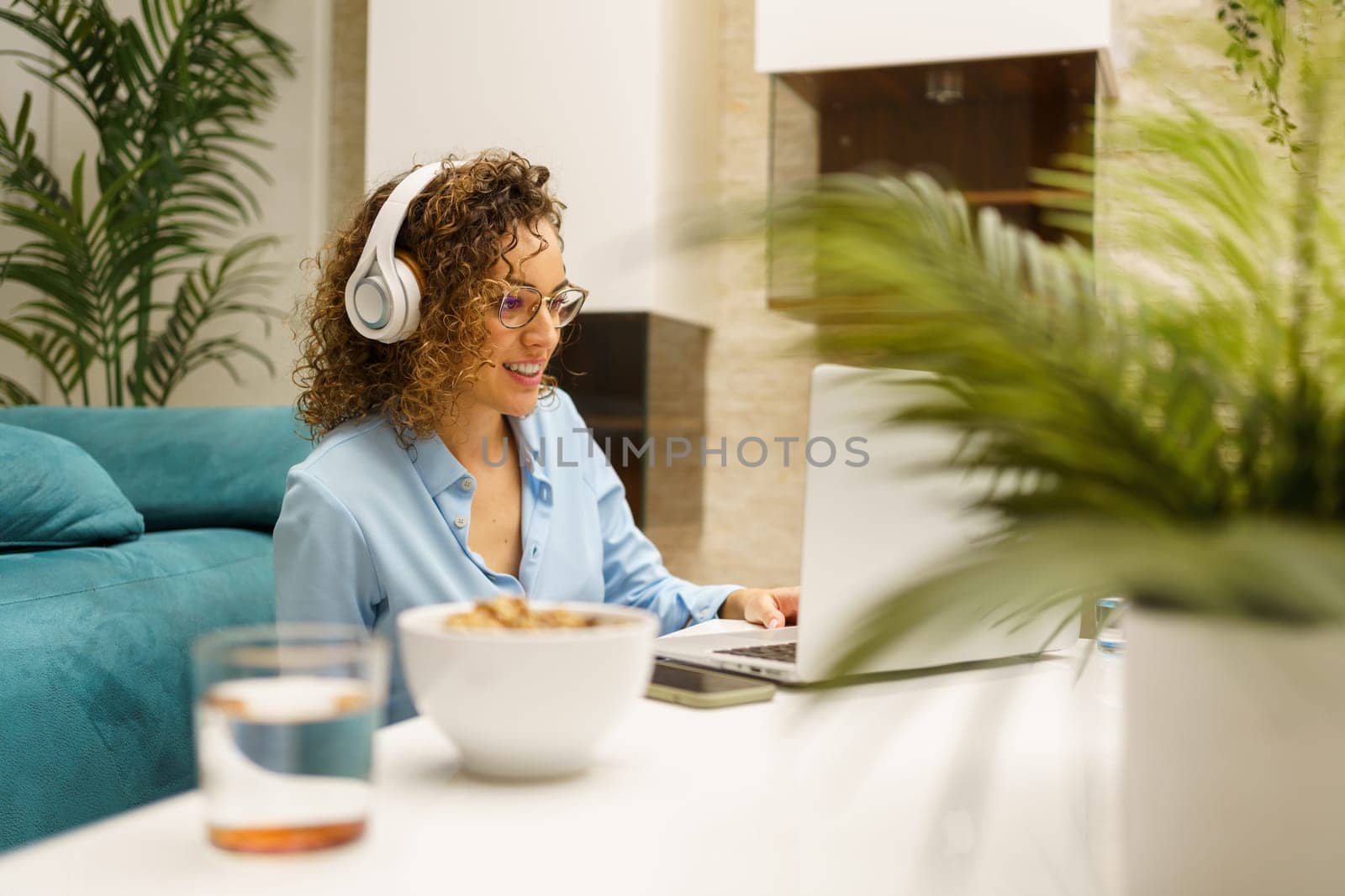 Young woman in headphones using laptop while sitting by white table in room by javiindy