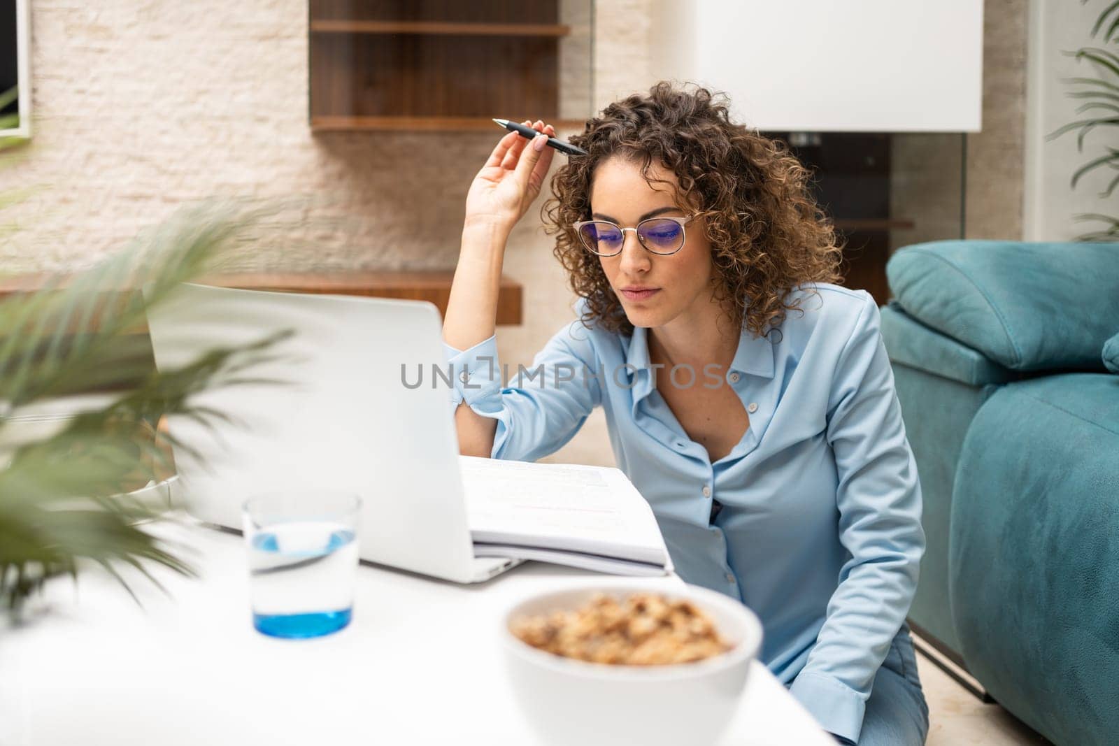 Focused young female freelancer sitting at table with laptop and notebook while working on project in living room at home