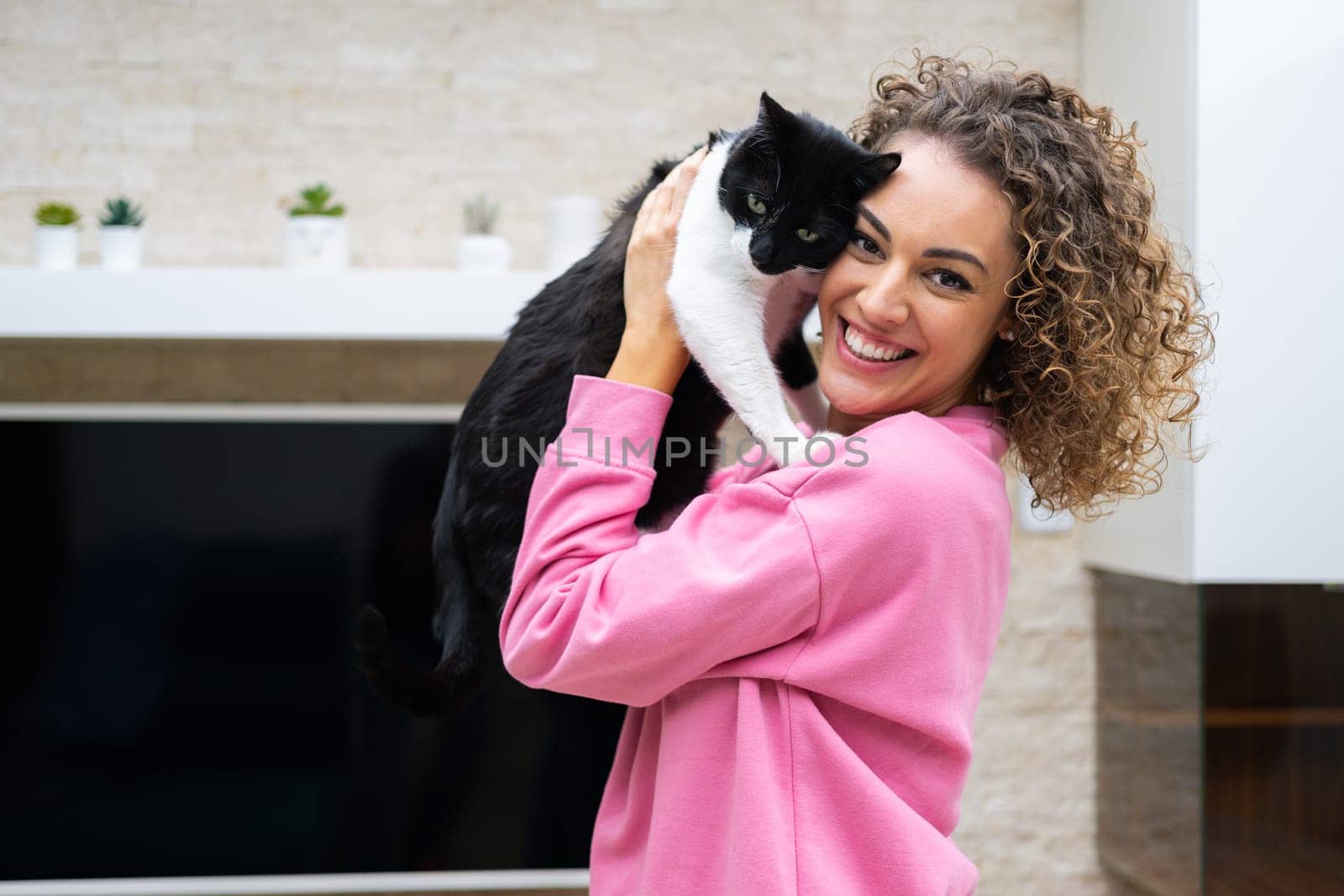 Side view of cheerful female with curly hair in casual cloth smiling and looking at camera while embracing cute cat in bright room