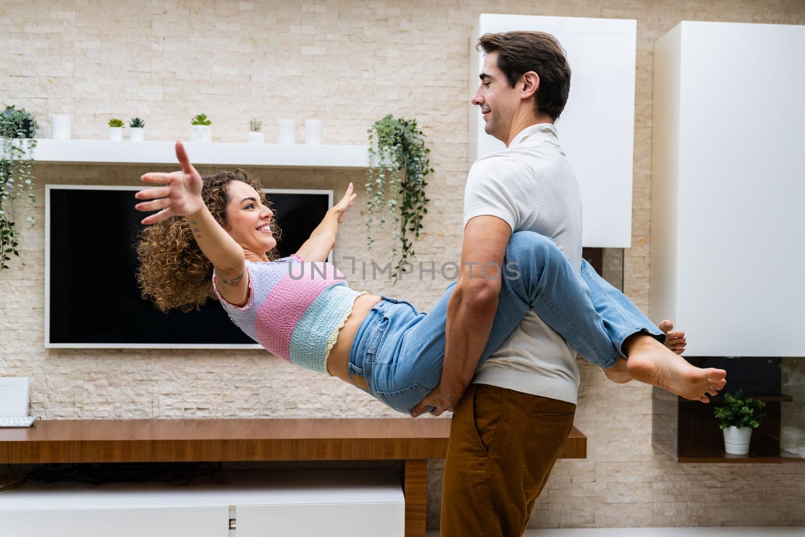 Side view of positive young couple having fun and enjoying time together while standing against led tv on wall with potted green plants at home