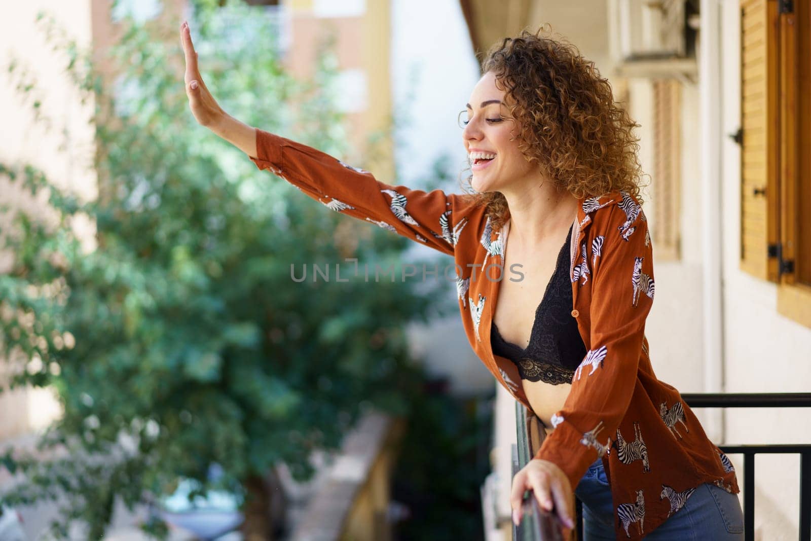 Side view of smiling curly haired female in stylish clothes standing on balcony and greeting with raised hand while looking down against blurred background