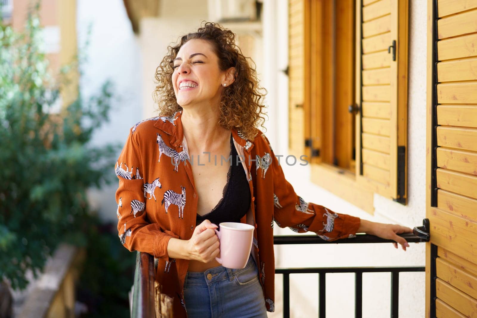 Cheerful young curly brunette haired female wearing stylish outfit with coffee cup leaning on balcony railing against blurred background