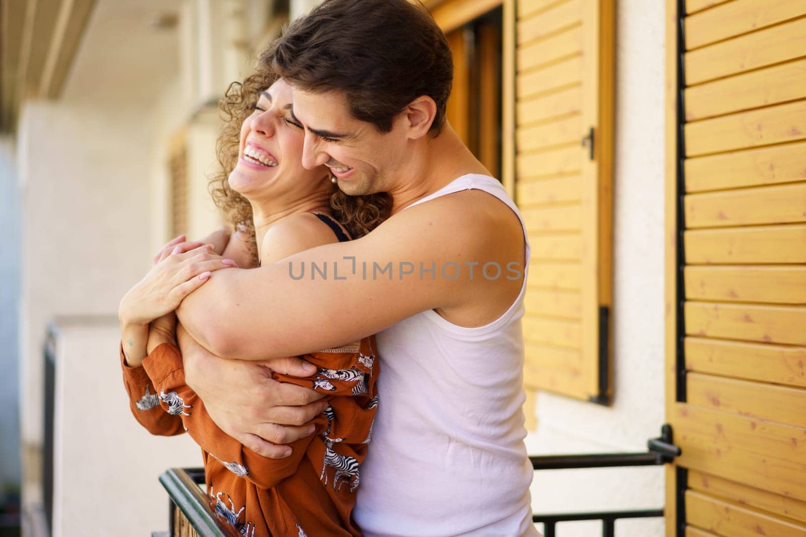 Side view of cheerful young couple in casual clothes, hugging and laughing together while standing by metallic handrail on balcony at facade of residential apartment on sunny day