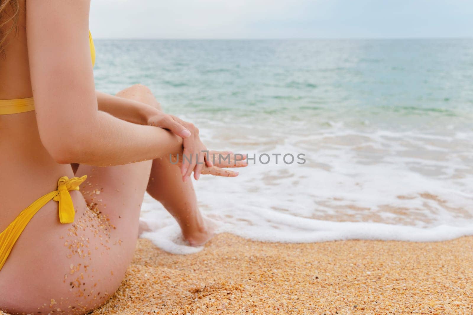 woman sits on the sand, touching her legs with her hand. a wave of sea water rolls over a woman's sexy body, an unrecognizable girl on vacation by yanik88