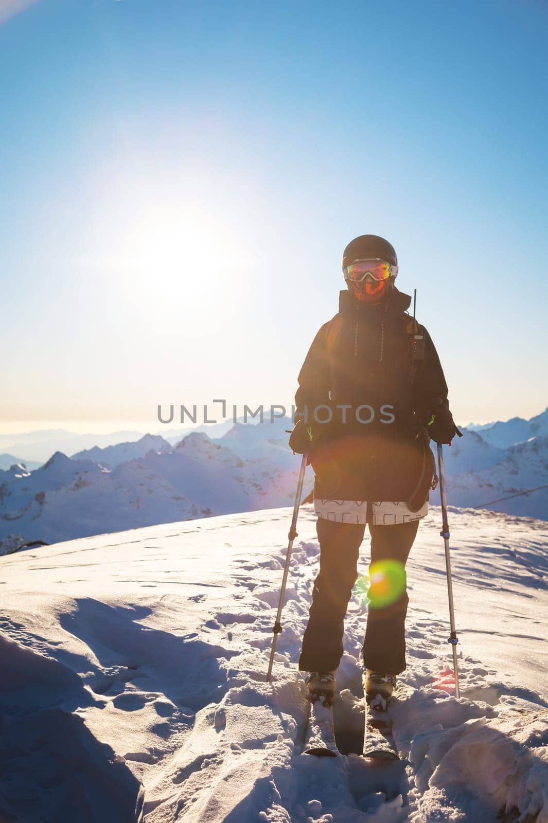 happy woman in a ski suit and helmet stands in the mountains against the backdrop of a mountain range on a sunny winter day. a moment of rest, relaxation, pleasure, winter holidays by yanik88
