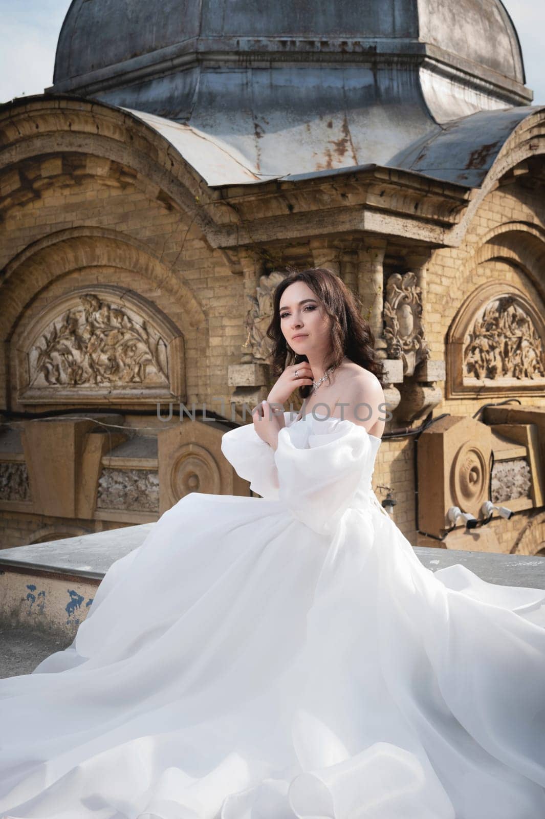A bride in a white dress sits on a ledge with an old building in the background. Young Caucasian woman straightens her hair at a wedding in an ancient city.