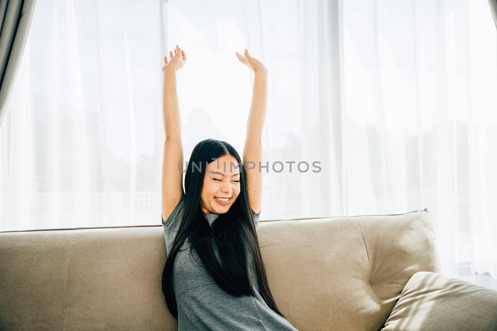 Smiling woman in luxury living room raises arms stretches after sitting. Lifestyle of a cheerful entrepreneur on sofa. Wellbeing joy and carefree relaxation.