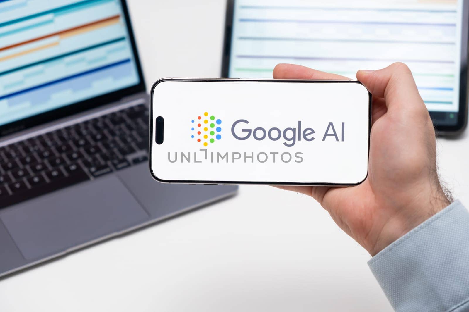 Google AI logo on the screen of a smartphone in mans hand with laptop and tablet with graphical diagrams by vladimka