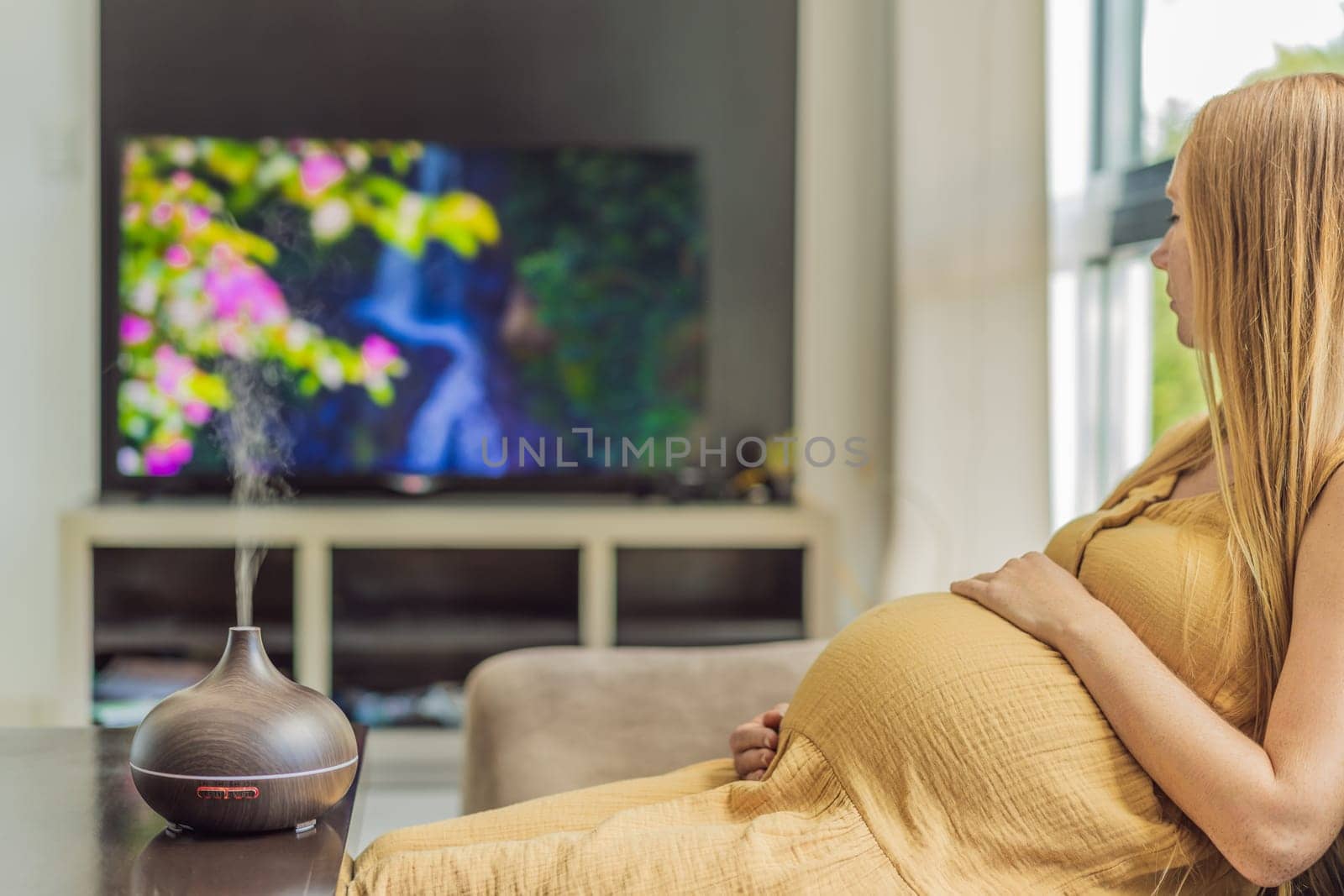 A blissful pregnant woman immerses in relaxation, savoring the soothing aroma from a diffuser while indulging in a calming TV video, embracing tranquility during her pregnant journey.