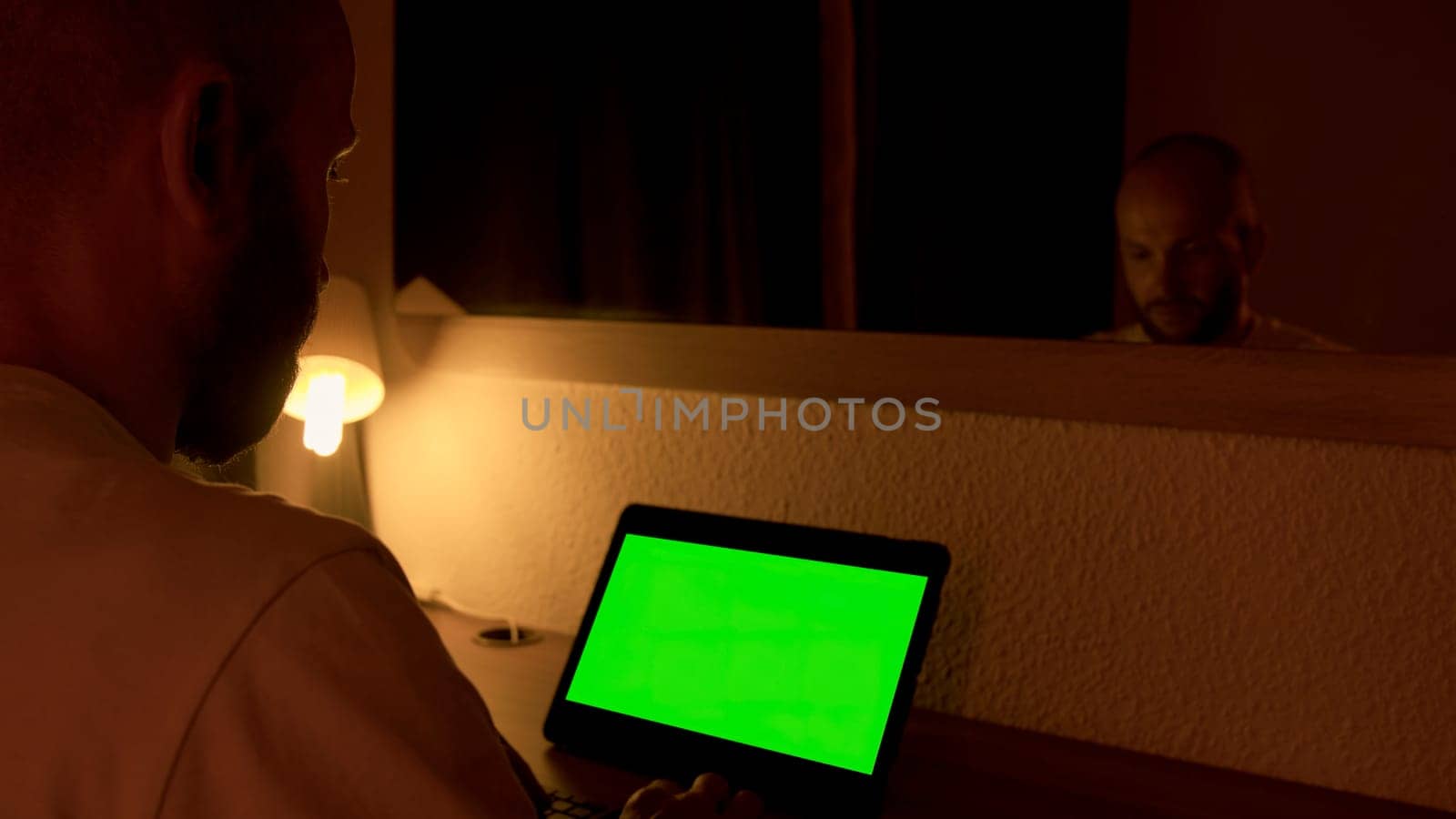 Man sitting at his desk, works on a laptop with green chroma key screen. Media. Man uses portable computer for work at home at night in front of mirror. by Mediawhalestock