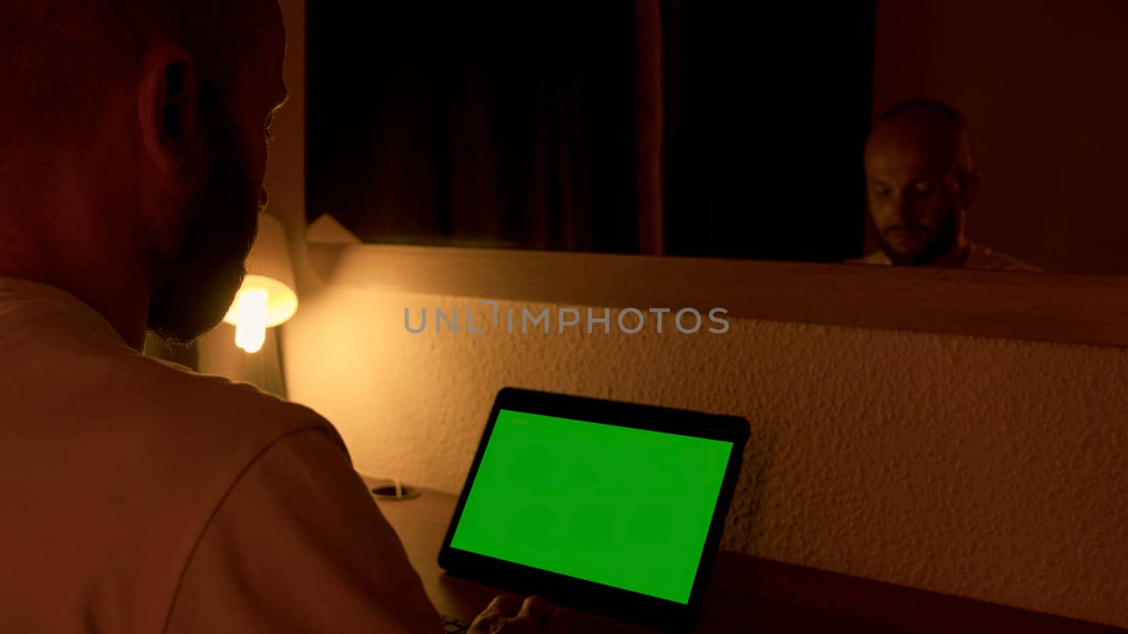View over the shoulder of a businessman working on laptop with green chroma key screen. Media. Young man working at the computer late at night in front of a mirror