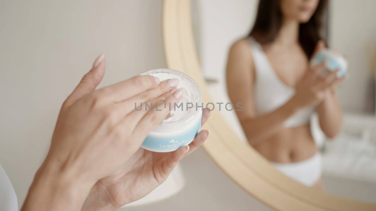 Women 's rest . Action . A beautiful young woman model in underwear who puts white cream on her body and poses next to a mirror. by Mediawhalestock