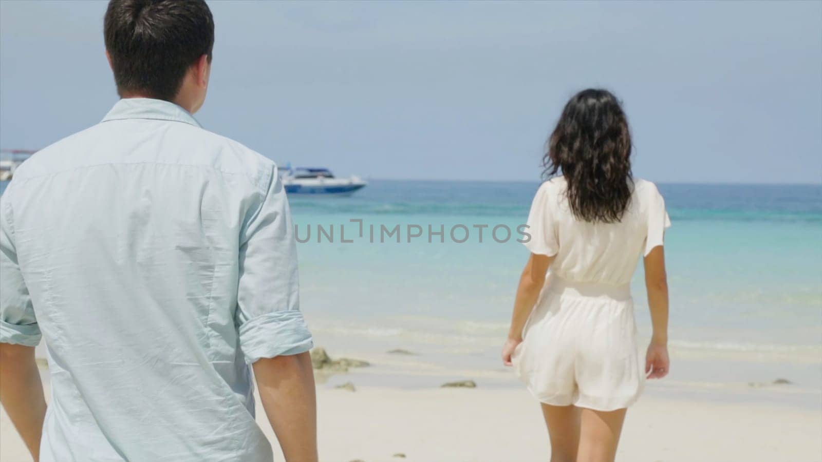 Cute couple in white clothing goes hand beach. Couple of teenagers running and flirting on the beach shore near the water. Woman walking on romantic honeymoon beach holidays holding hand of boyfriend following her, view from behind. A young girl holding hands on the beach.