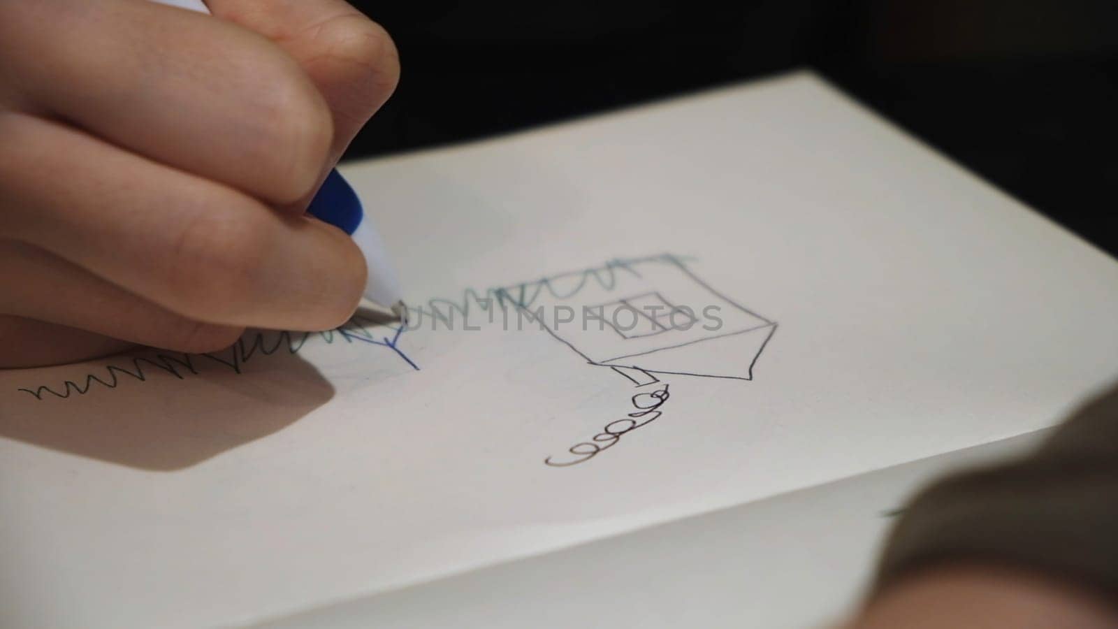 Child drawing a picture of his family and a gouse in notebook, close up. Small kid using a pen to draw a sketch of his mom and dad near the house.