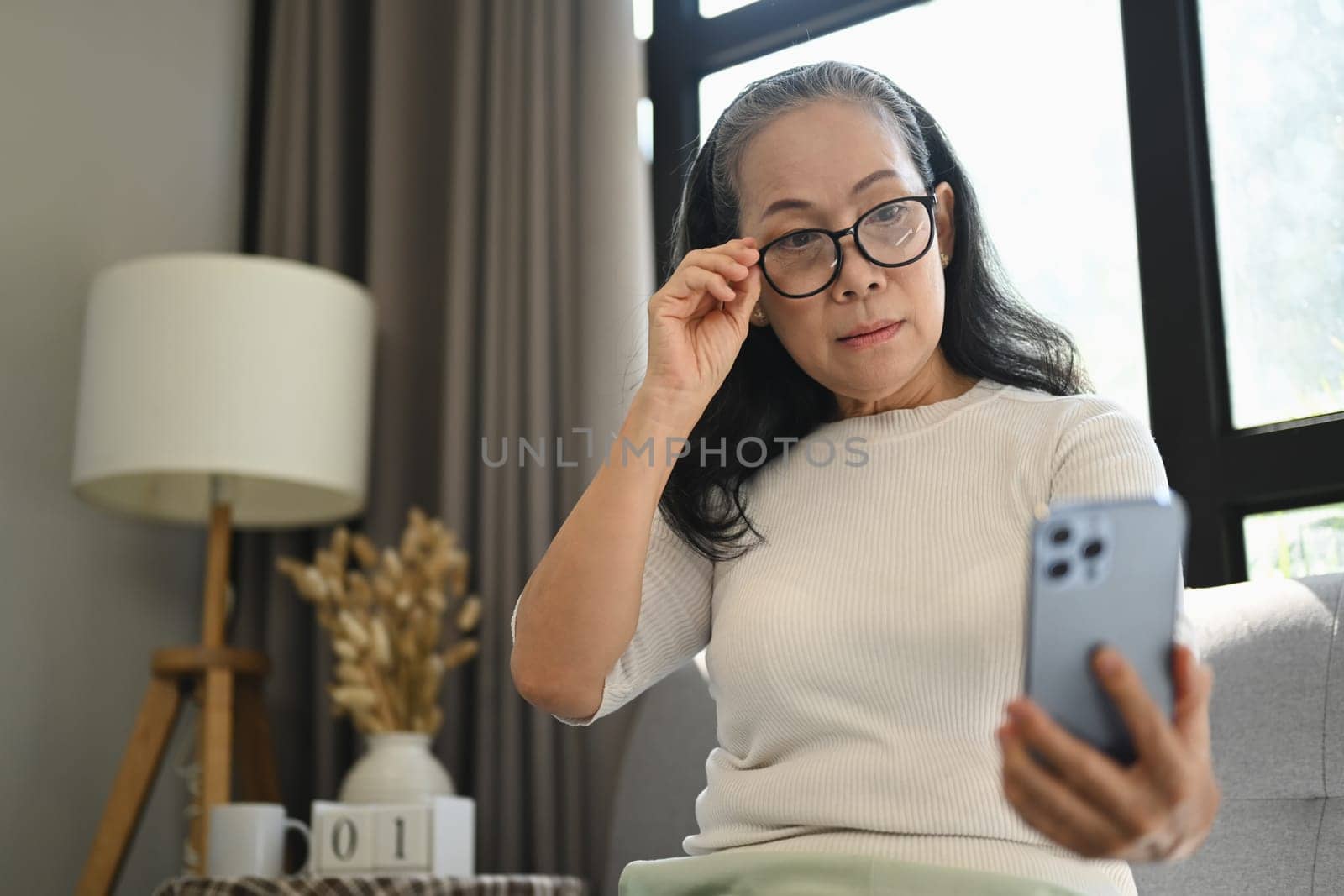 Beautiful middle age woman in glasses checking apps on mobile phone while sitting on couch.