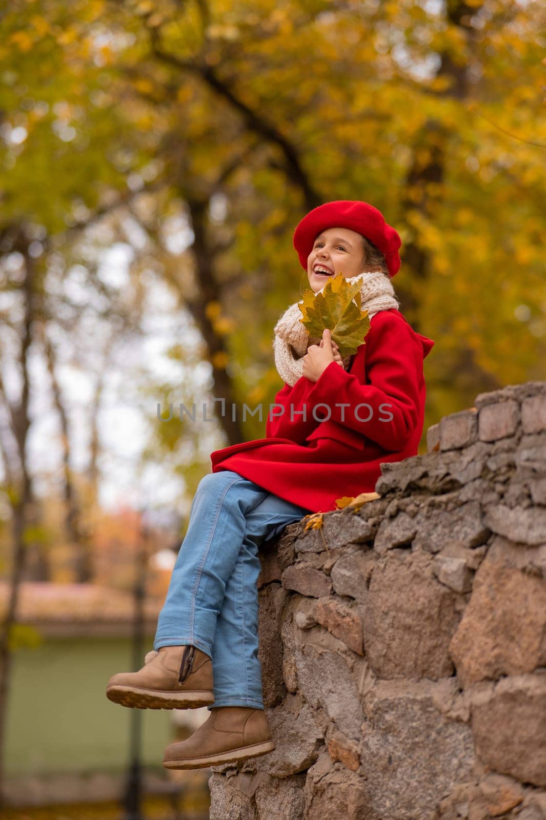 Smiling caucasian girl in a red coat and beret sits on a brick wall on a walk in autumn