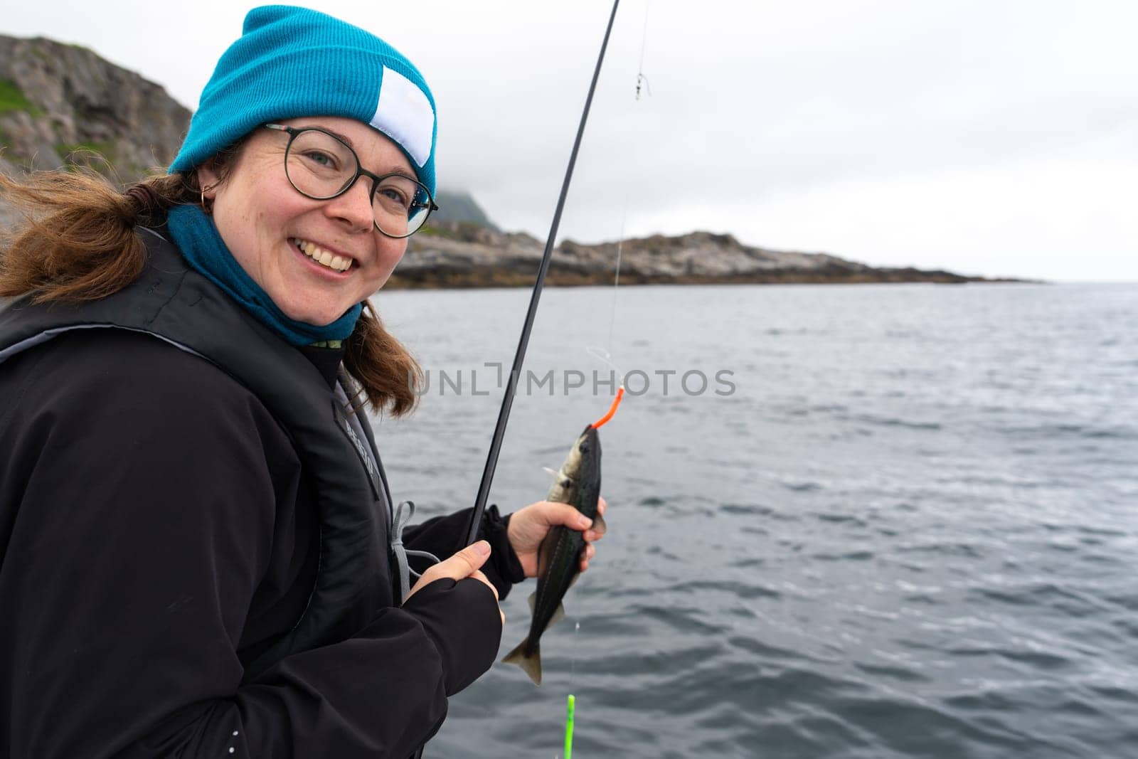 A smiling female fisherman proudly displays a large catch while standing on a boat in the Norwegian Sea, enjoying her hobby and the beauty of the ocean on a sunny day.