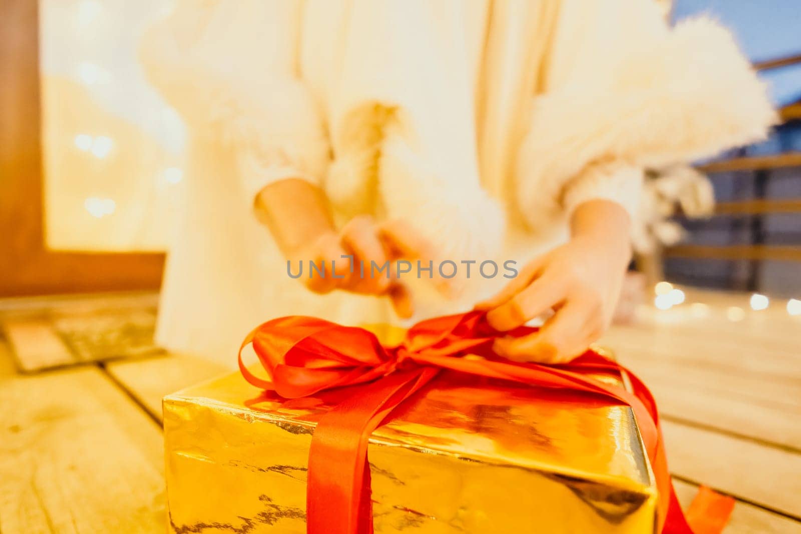 A woman in a white dress is holding a gold box with a red ribbon. She is wearing a crown on her head. The scene takes place in a room with a door and a window. The woman appears to be opening the gift box.