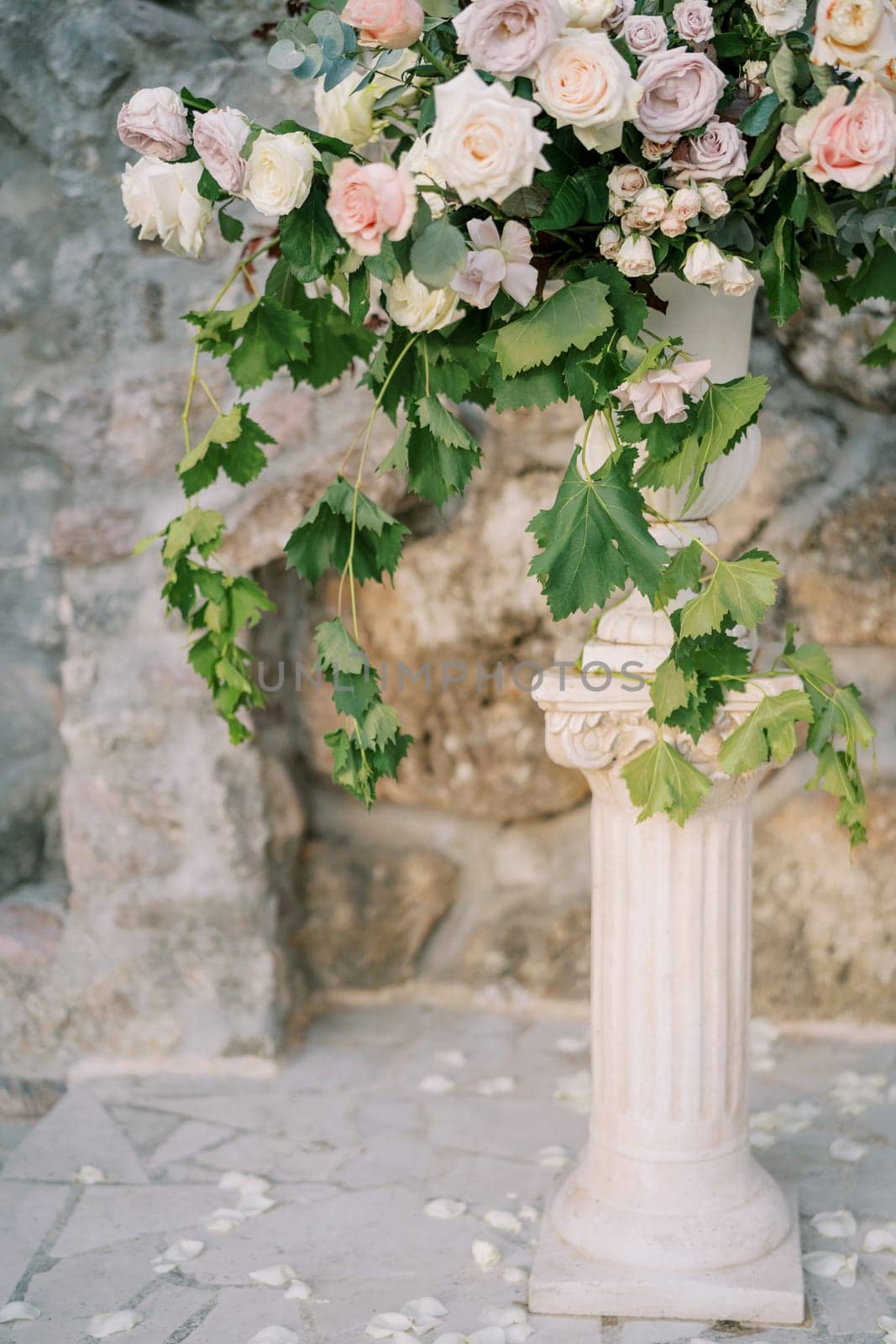 Lush bouquet of flowers stands in a vase on a pedestal near a stone wall in the garden by Nadtochiy