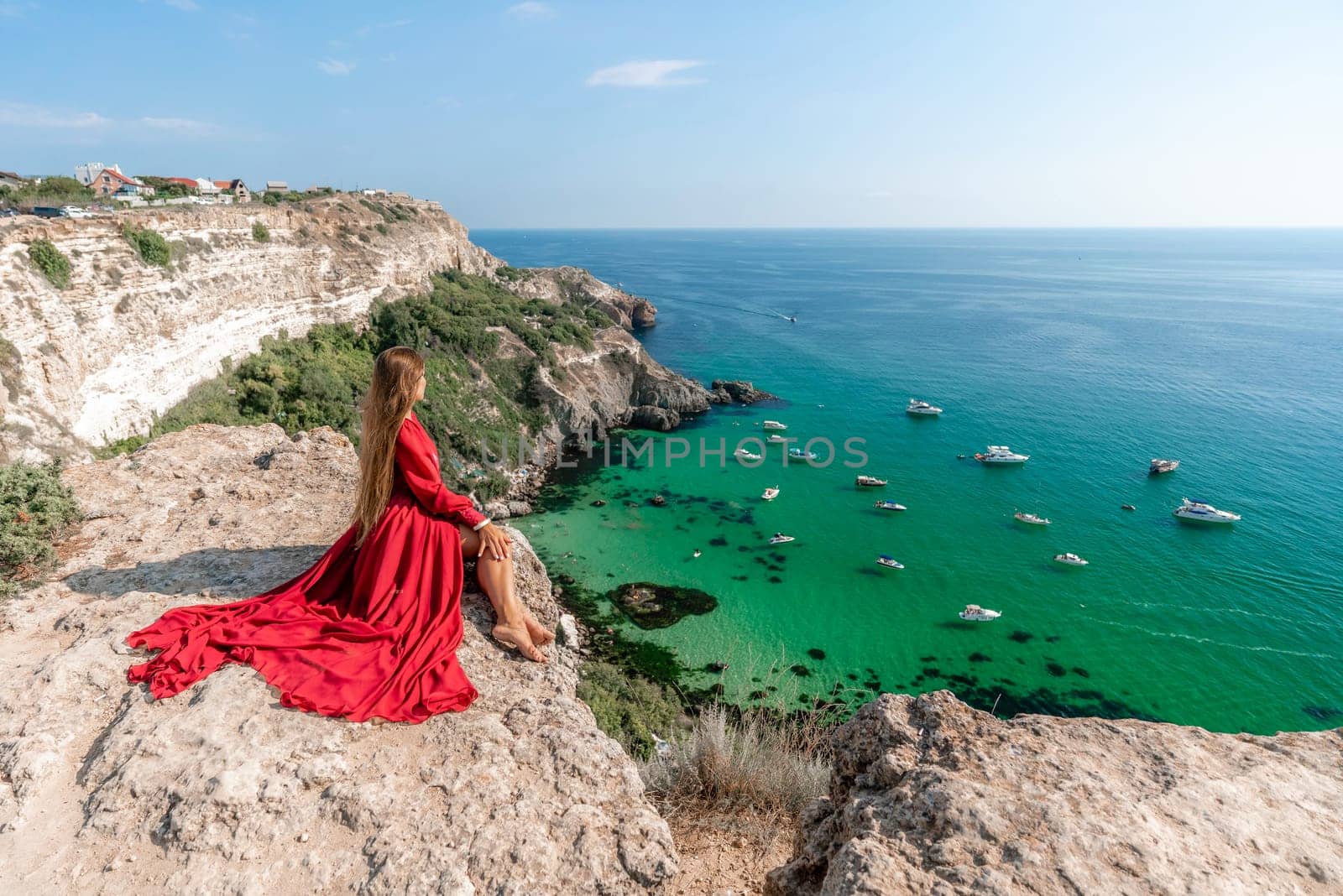 Woman red dress sea. Happy woman in a red dress and white bikini sitting on a rocky outcrop, gazing out at the sea with boats and yachts in the background
