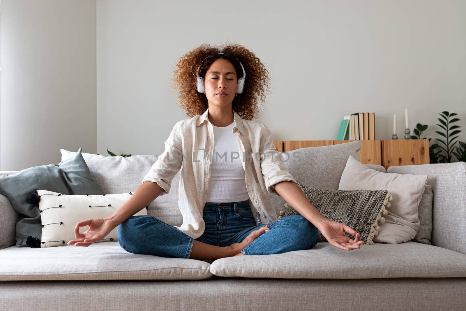 Front view of multiracial hispanic young woman meditating on the couch listening to guided meditation using wireless headphones. Technology and mental health concept.