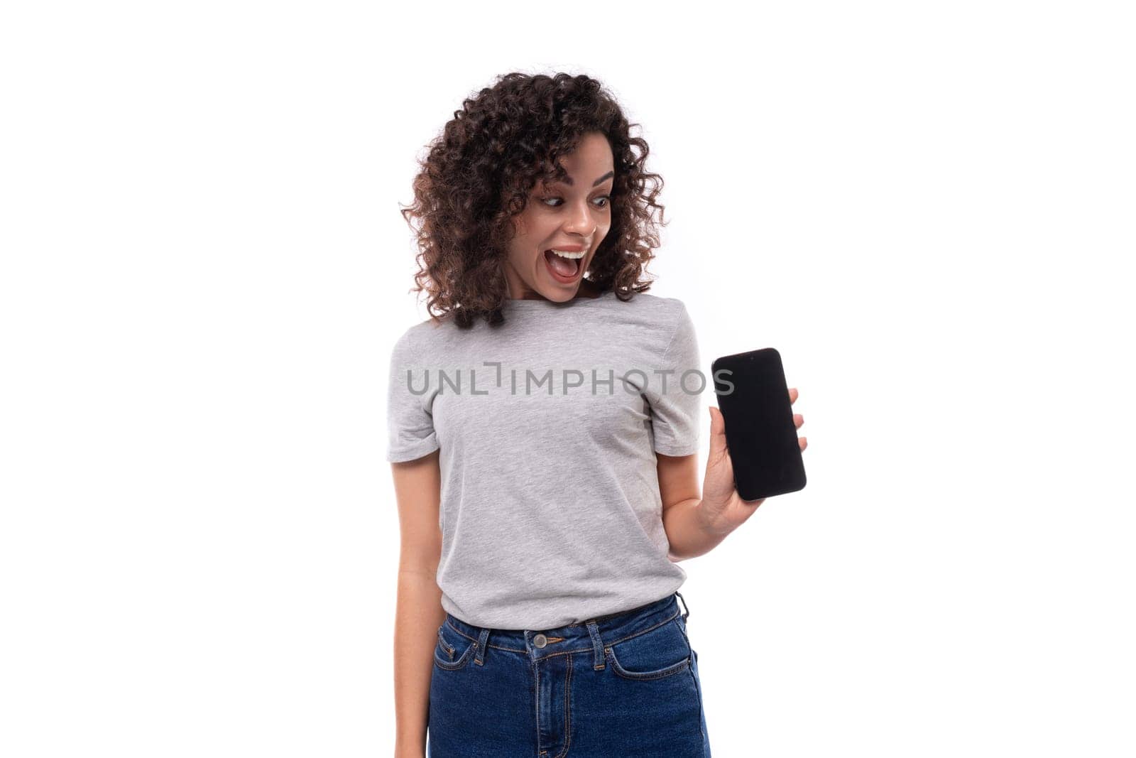 portrait of a young surprised joyful 30 year old caucasian woman in a gray t-shirt holding a smartphone with a mockup on a white background with copy space.