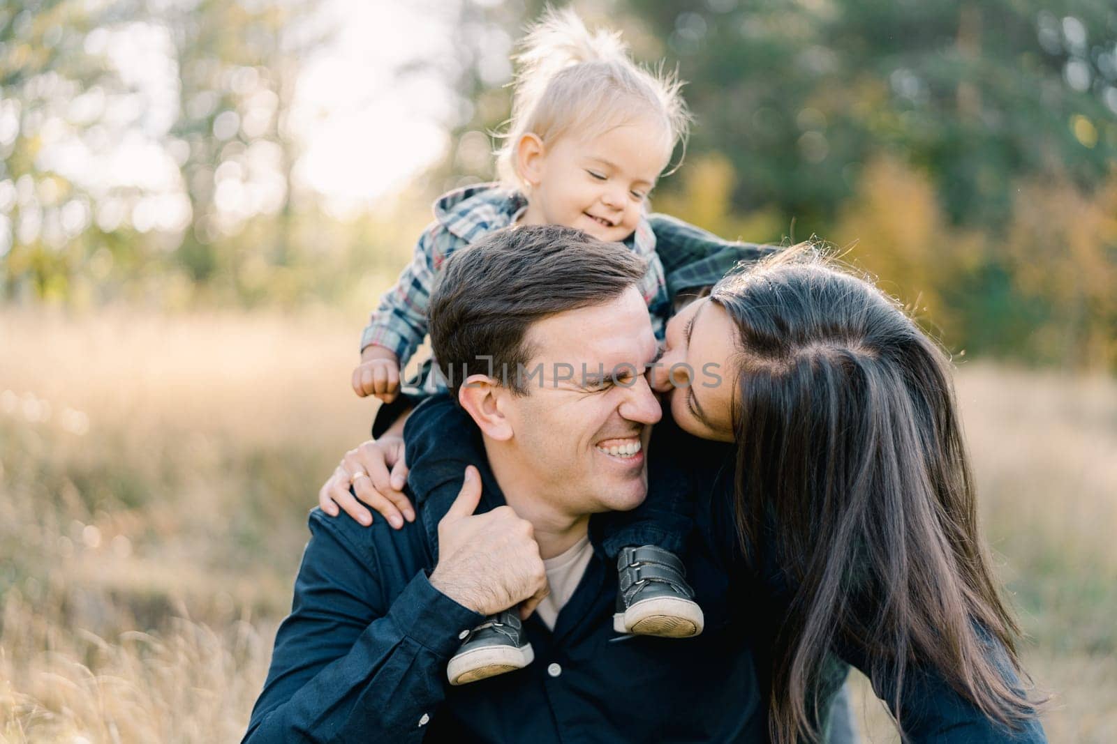 Mom hugs and kisses dad on the cheek from behind with a little girl on his shoulders. High quality photo