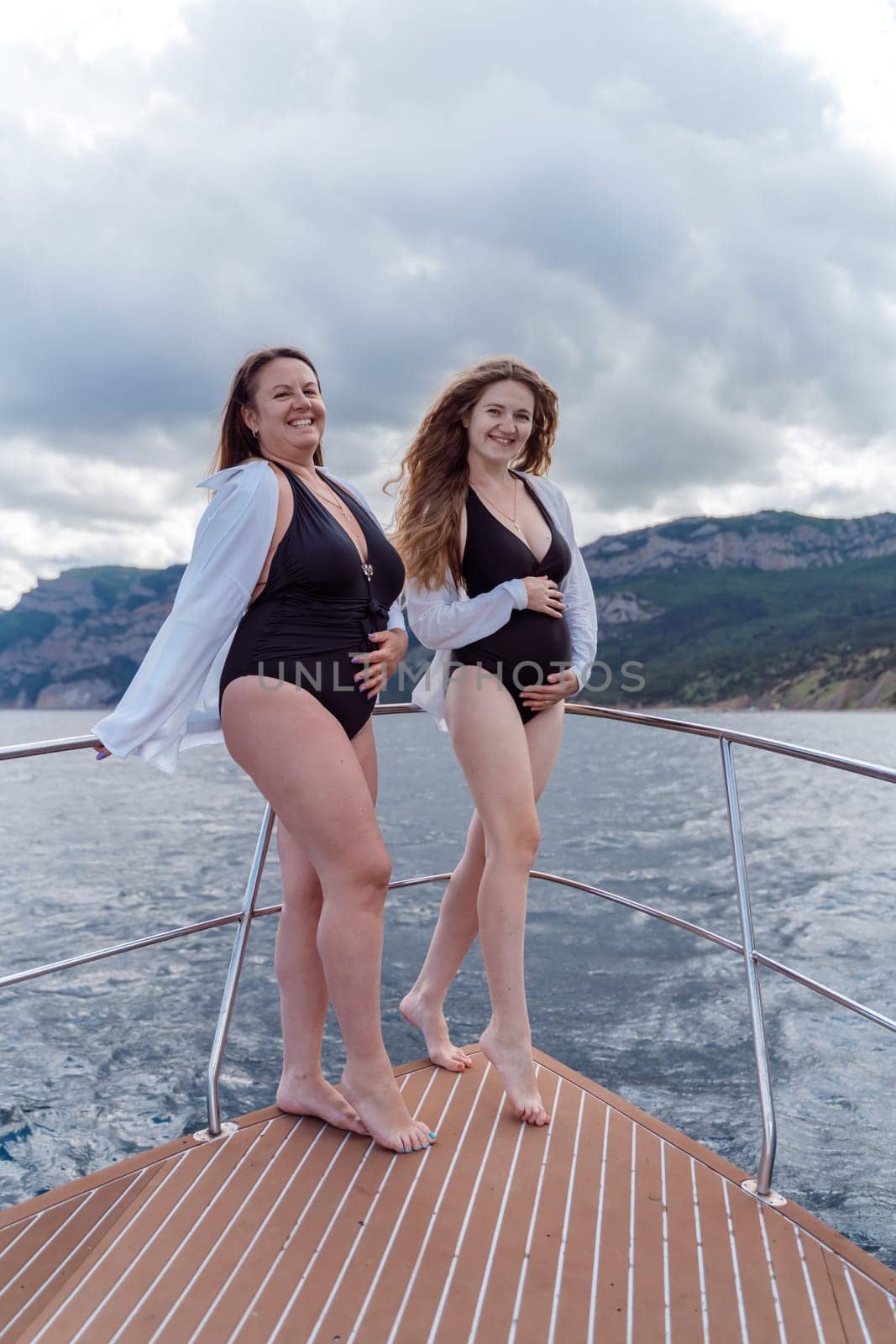Pregnant on a yacht. Happy models in a swimsuit posing on a yacht against sky with clouds and mountains