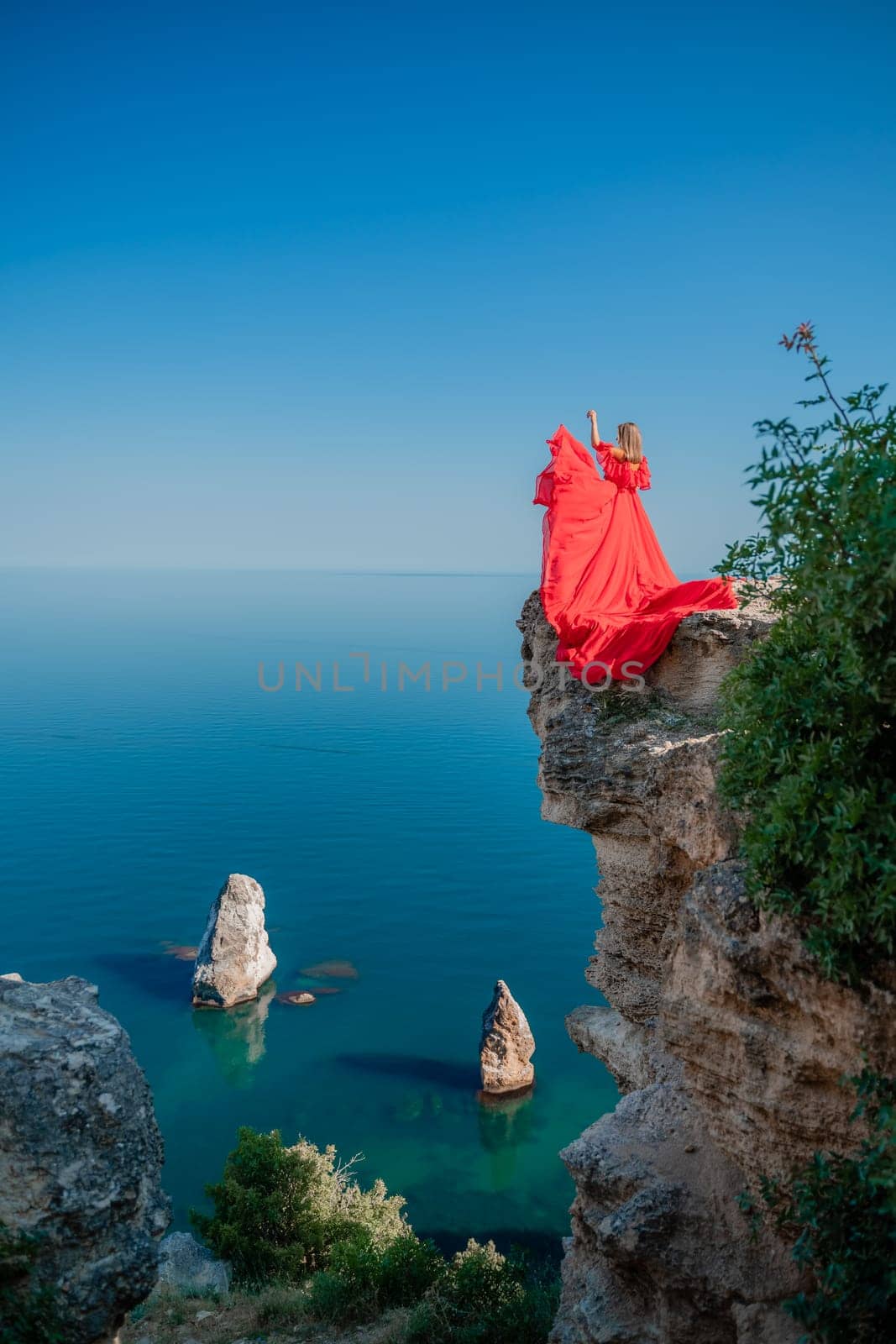 Red dress sea woman. Happy woman with flowing hair in a long flowing red dress stands on a rock near the sea. Travel concept, photo session at sea by Matiunina