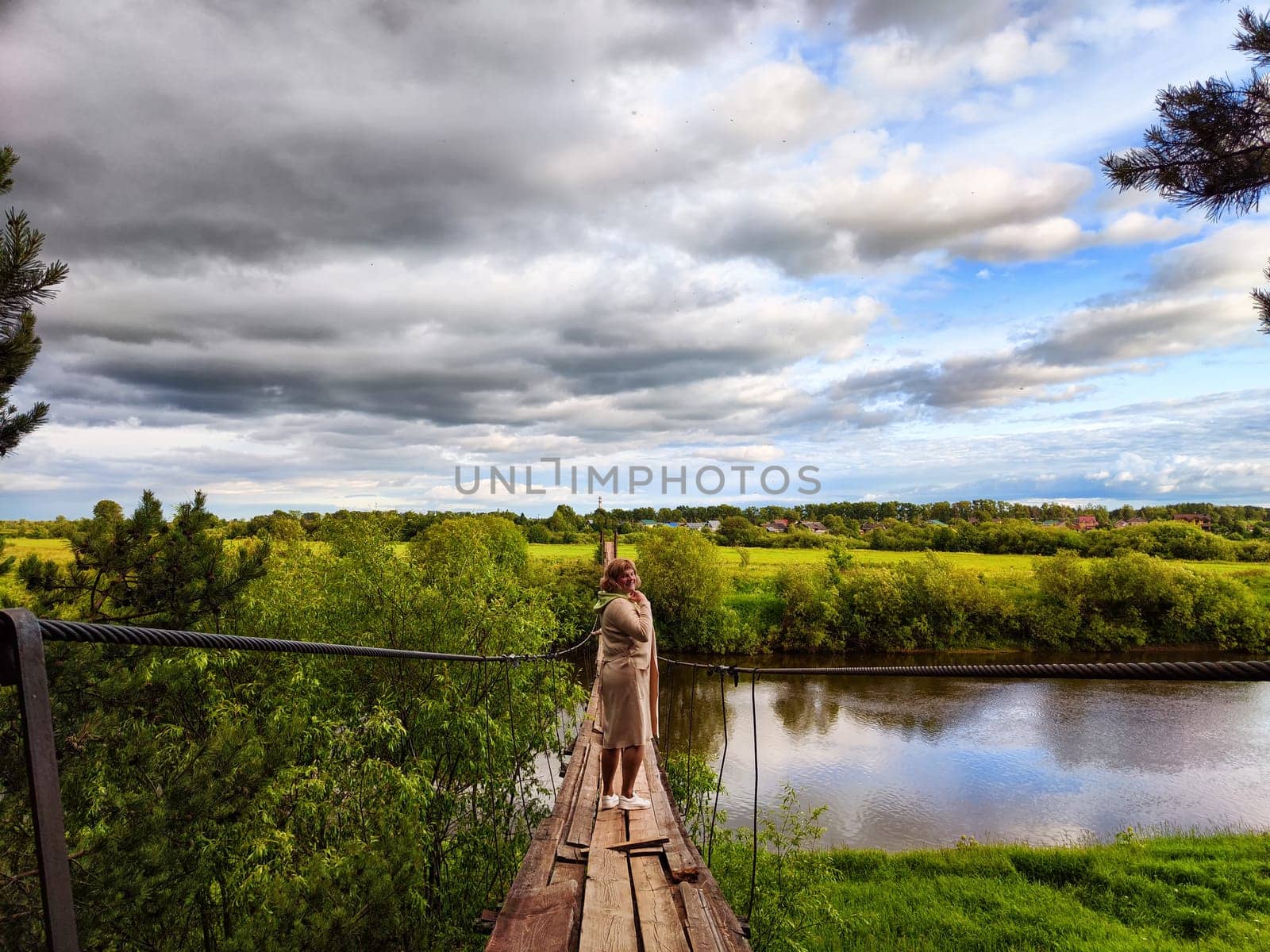 Tourist girl walks along an old wooden suspension bridge over the river and the sky with disturbing clouds. Suicide in nature. The concept of depression and hopelessness of life