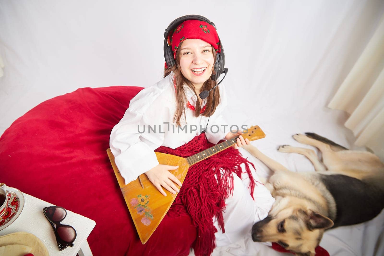 A girl in red scarf, white shirt, with shawl and balalaika in black sunglasses with a shepherd dog. A cheerful radio presenter with headphones and a microphone sings and has fun with an animal friend by keleny