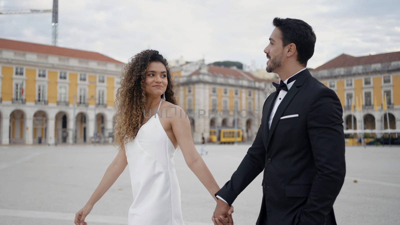 Young and beautiful couple walking along historic buildings and talking. Action. Man in suit and woman in white dress holding hands in the city square. by Mediawhalestock