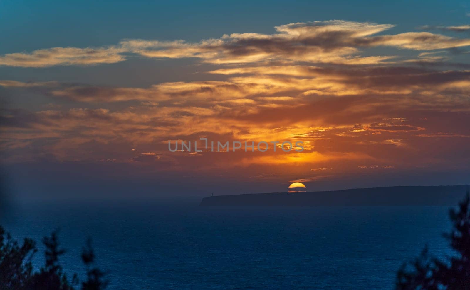 A sunset illuminates an endless sea as a lighthouse stands atop cliffs.