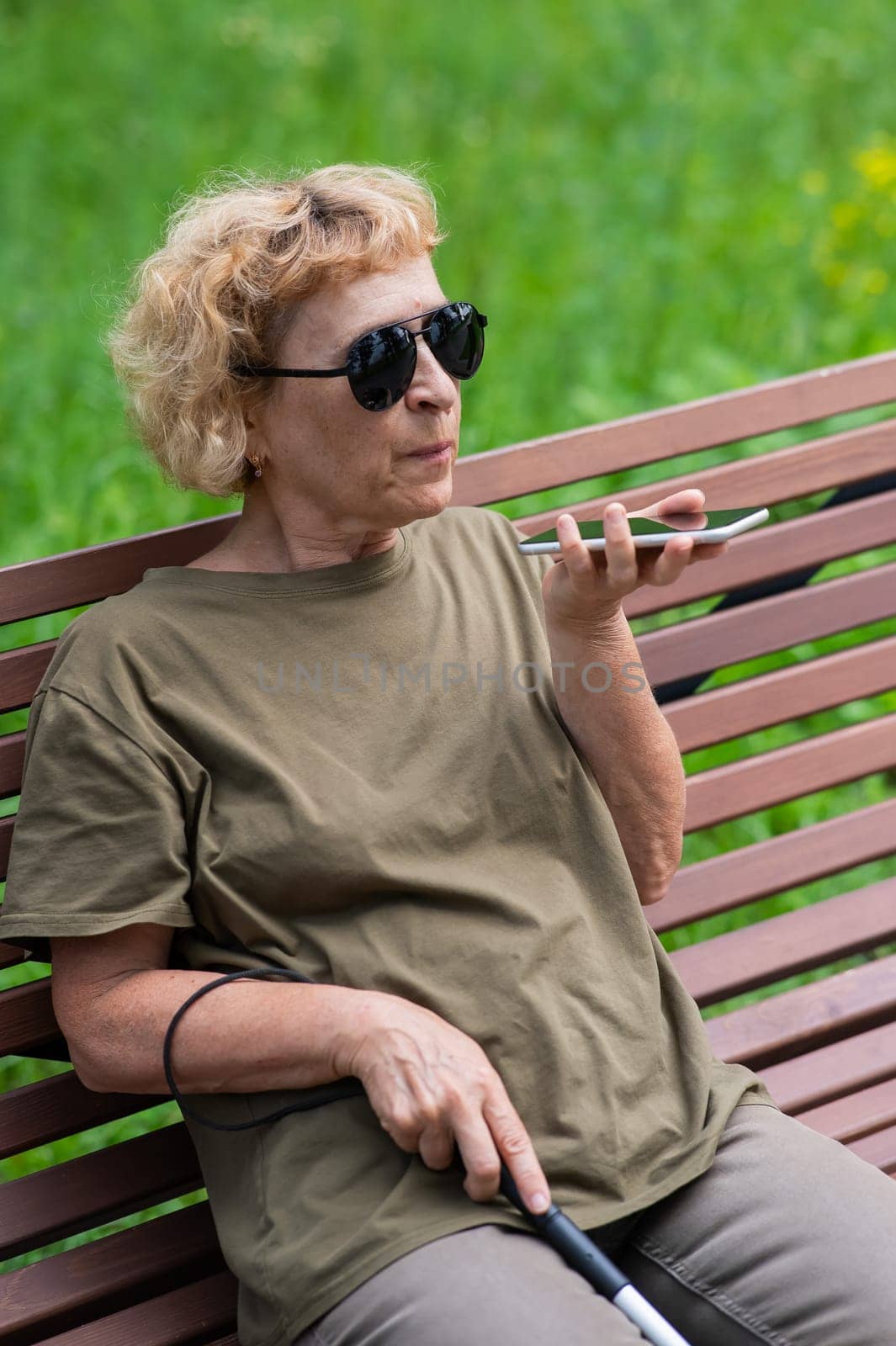 An elderly blind woman sits on a bench in the park and talks on a smartphone. Vertical photo. by mrwed54