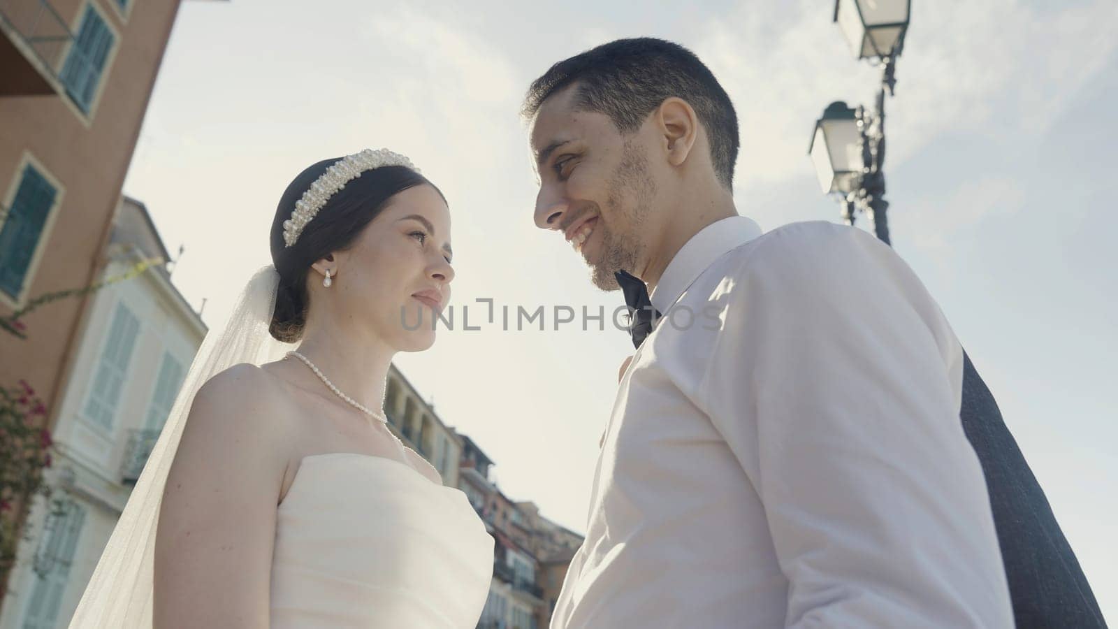 Bride and groom walking in a city street in summer day. Action. Low angle view of a woman with decoration on her hair and a veil standing near her groom in white shirt against the sun. by Mediawhalestock