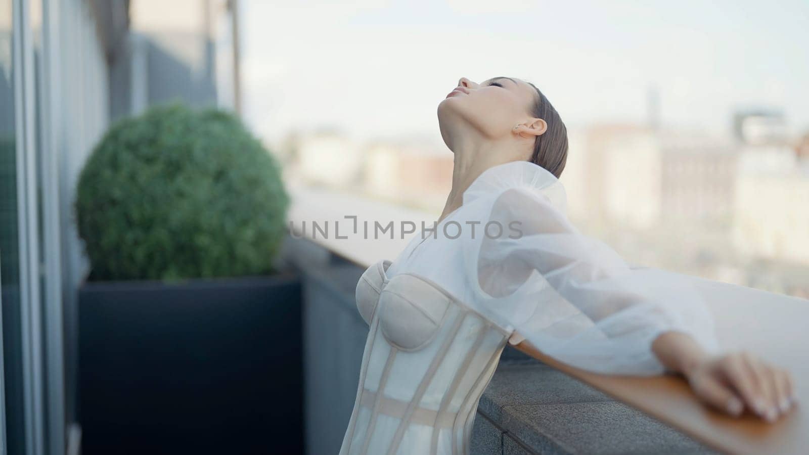 Young beautiful bride in a white wedding dress standing on a balcony on blurred city background. Action. Elegant woman with the waving silk scar tied around her ponytail