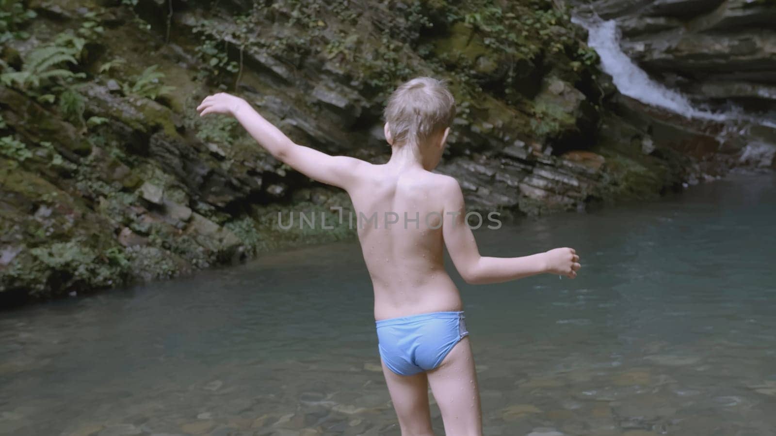 The boy enters the water near the waterfall to swim. CREATIVE. A white child in blue shorts enters the water. In the mountains there is a guy waist-deep in water.