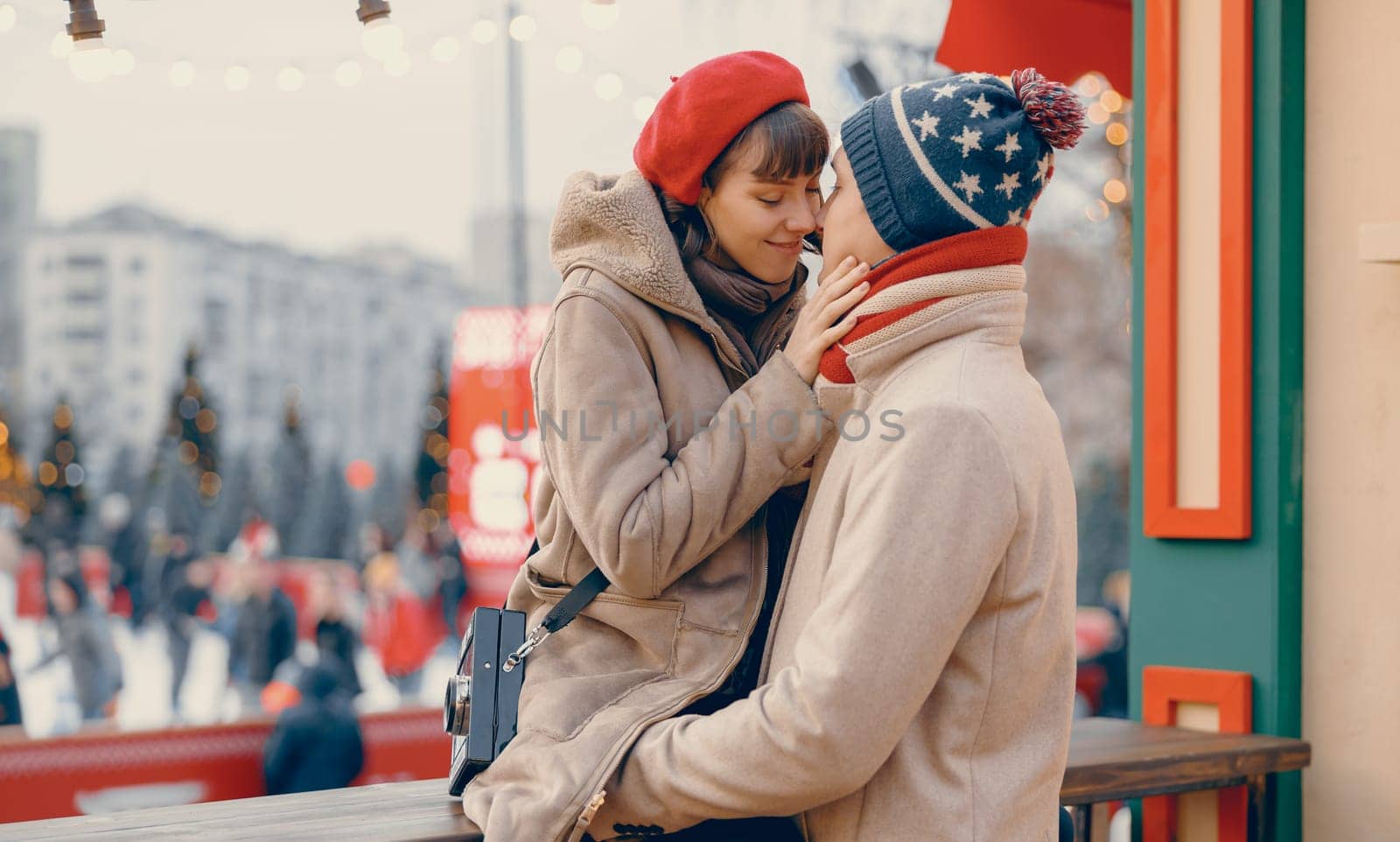 A woman tenderly touches her partner's face, sharing a close moment at a Christmas market with festive lights twinkling in the background.