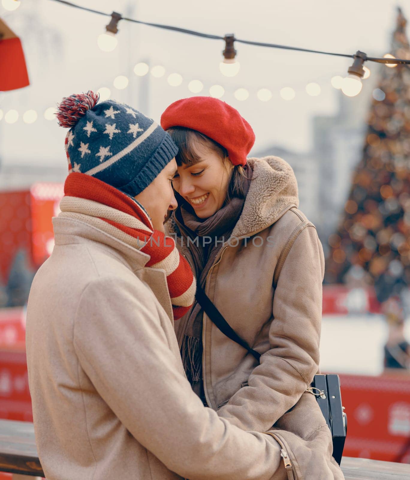 A romantic scene unfolds with a couple kissing gently amidst the twinkling lights of a holiday market, their winter attire adding warmth to the moment