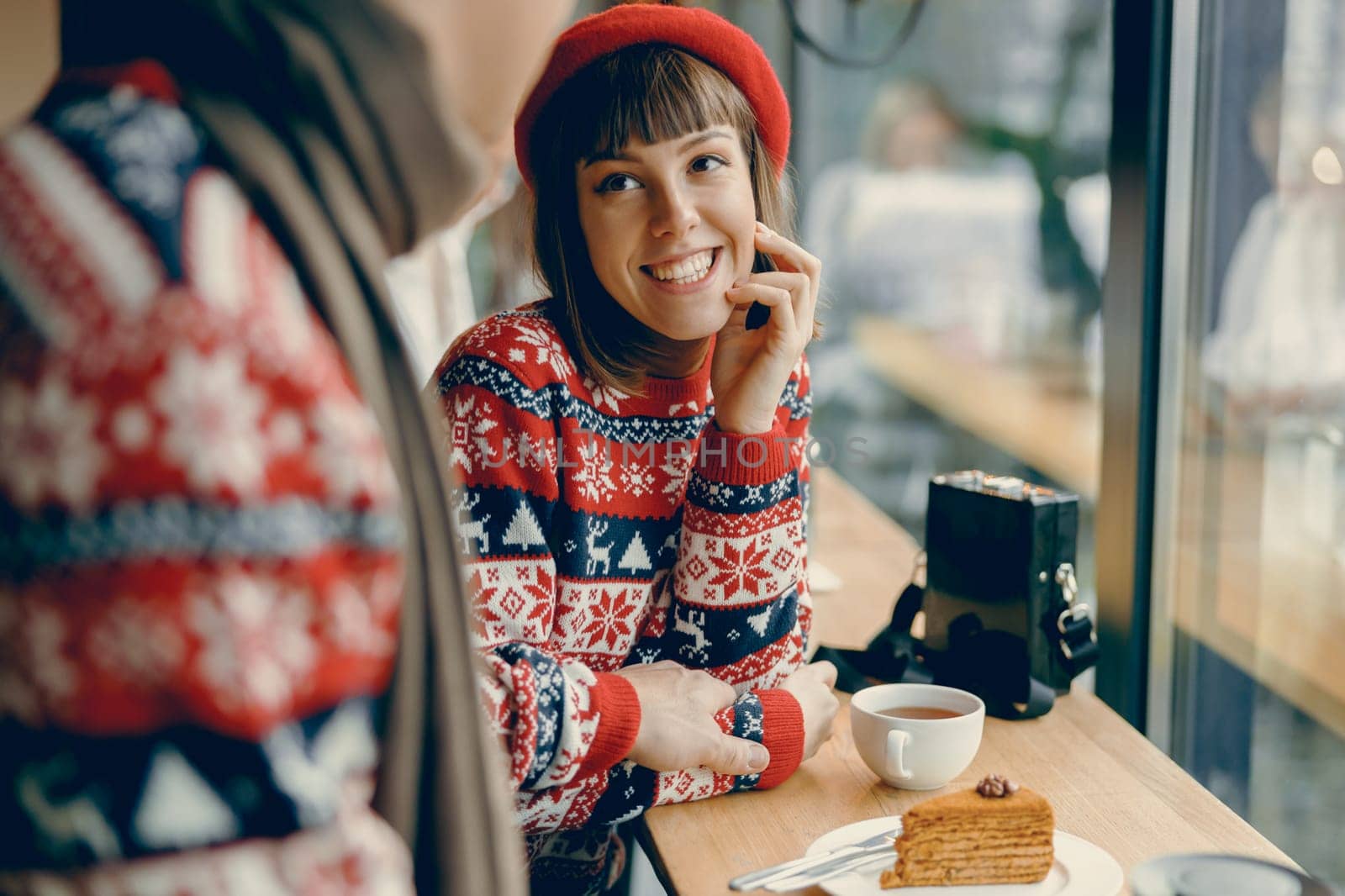 Smiling brightly, a woman in a holiday-themed sweater and red beret enjoys a cozy coffee date, exuding winter cheer