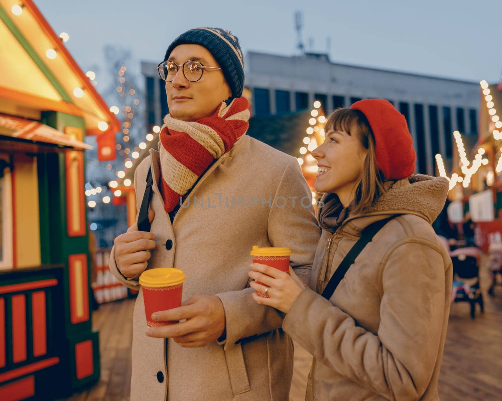 A couple stands close, holding warm drinks in a brightly lit Christmas market, with the woman looking up at her partner in a moment of shared contentment