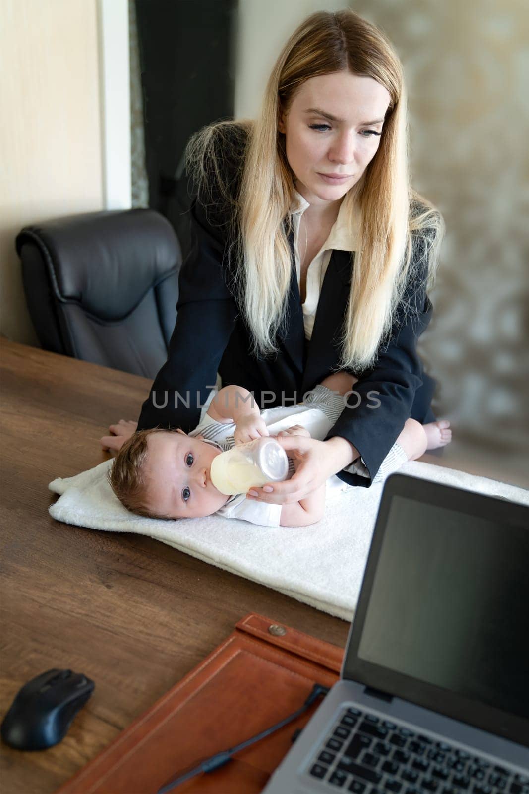 Young business woman feeds baby from bottle on desktop in office, combining it with work on laptop, vertical frame