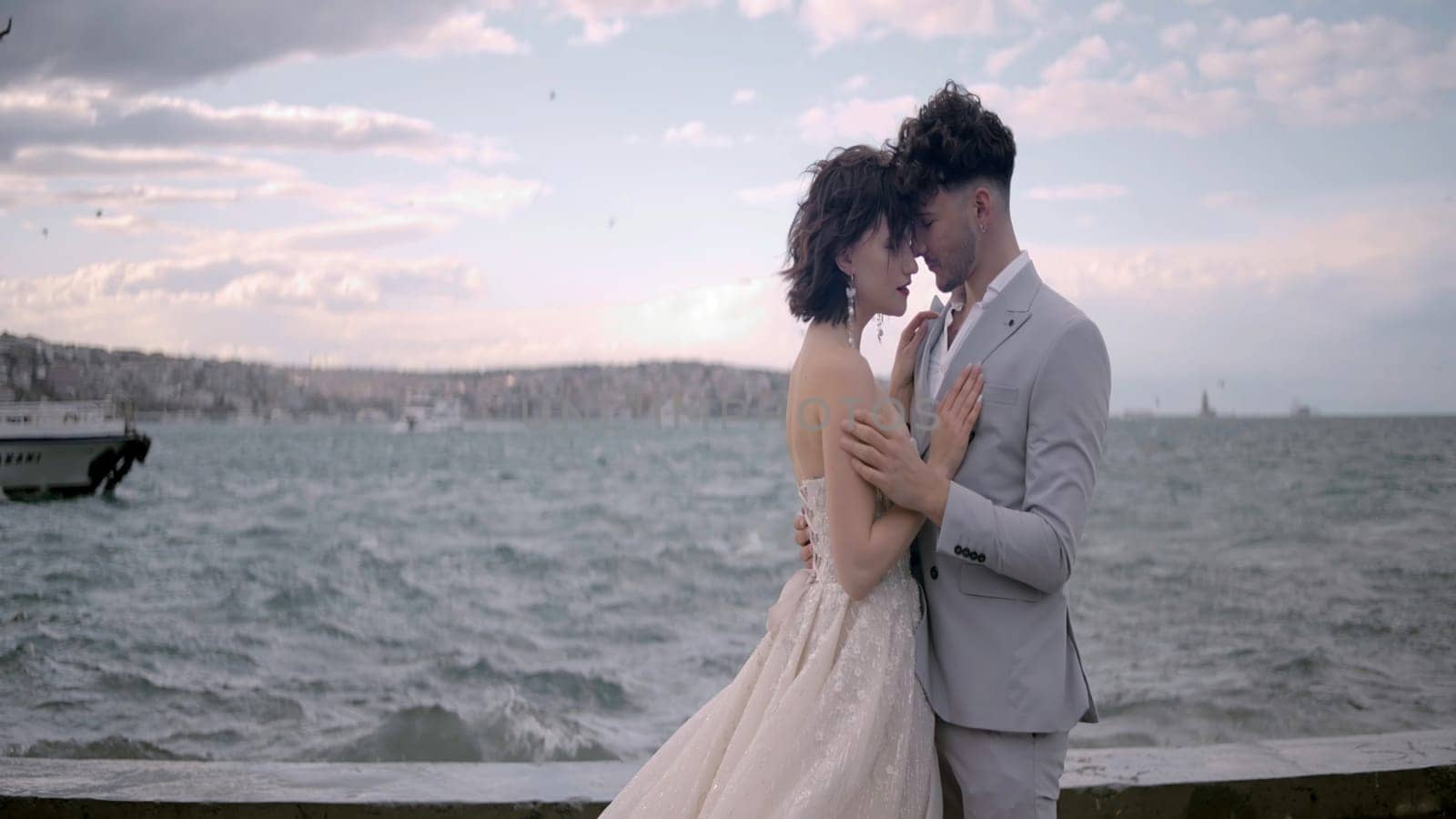 Newlyweds on background of waves. Action. Beautiful newlyweds are standing on pier on background of sea in windy weather. Newlyweds hug on background of surge of waves at pier.