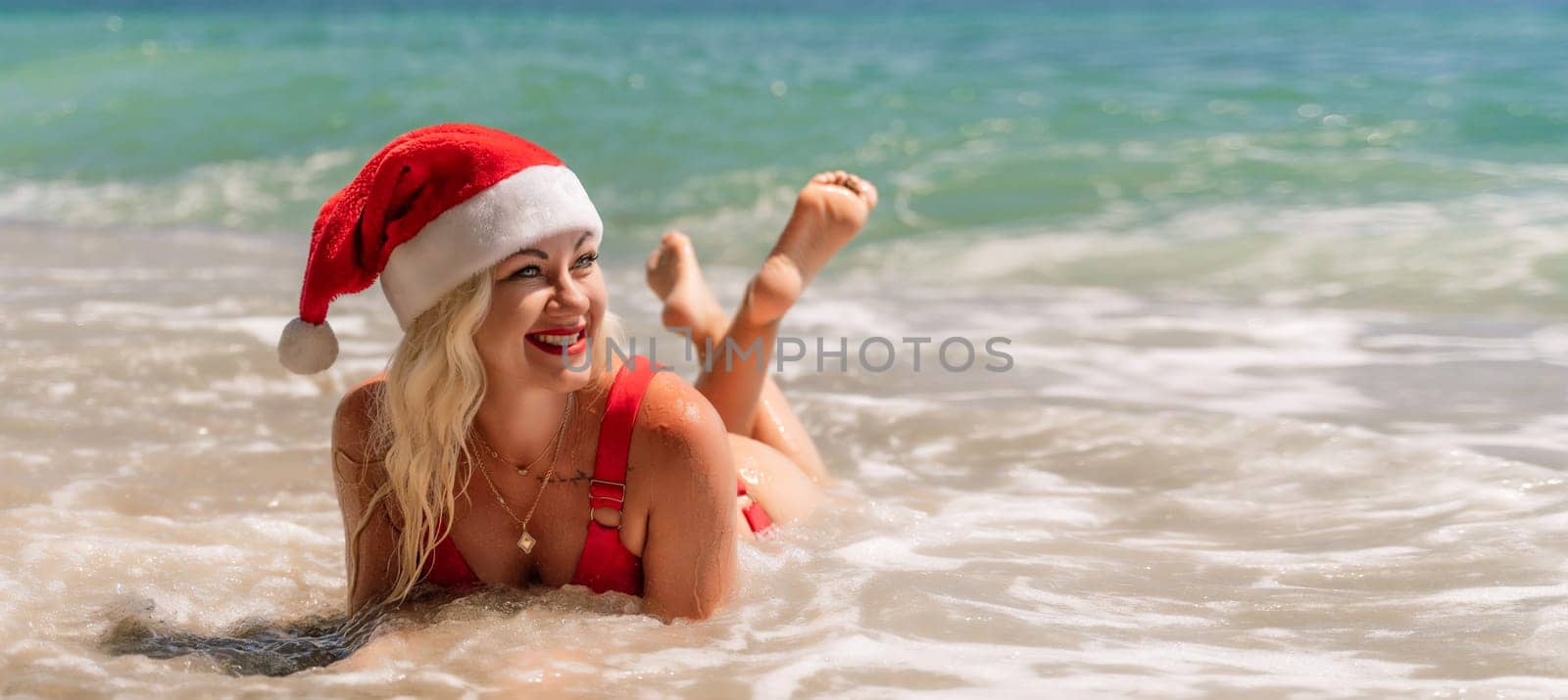 Female beach Santa hat wave coast. beach relaxation seaside. A woman in a red swimsuit enjoying her time on the beach, lying on the sand and being covered by a wave. by Matiunina