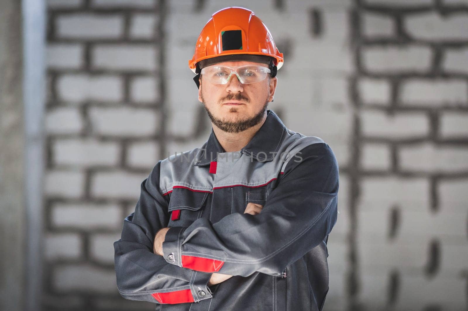 Portrait of a serious bearded Caucasian worker in overalls, safety helmet and safety glasses at a construction site.