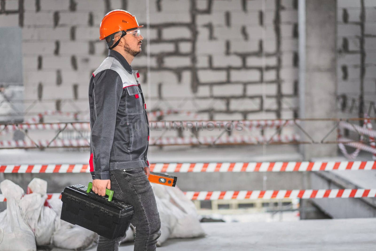 A worker in overalls and a helmet came to a new work site. Builder in uniform carrying toolbox at construction site, copy space by Rom4ek