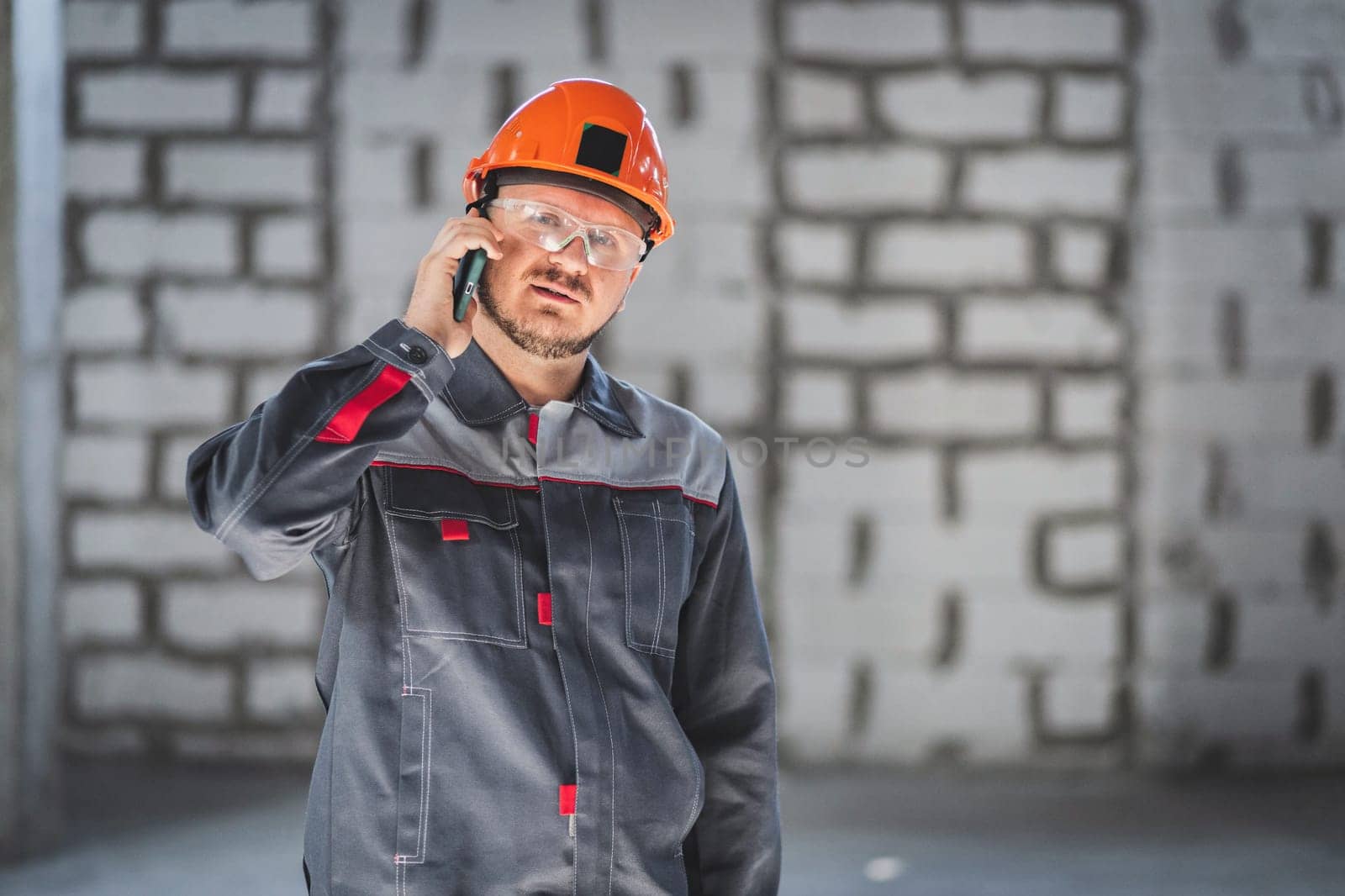 Bearded construction caucasian worker in boilersuit, helmet and protective glusses talking on a smartphone at a construction site by Rom4ek
